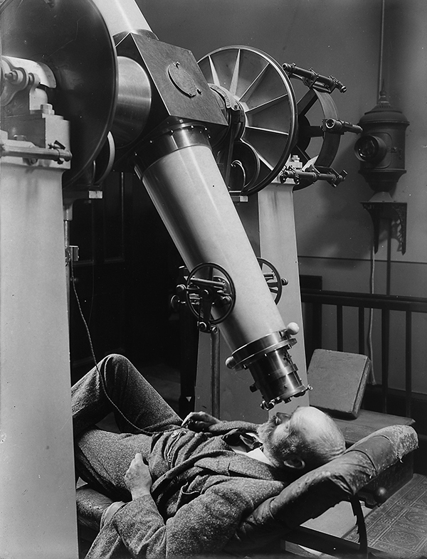black and white image of a reclined man looking through a large telescope